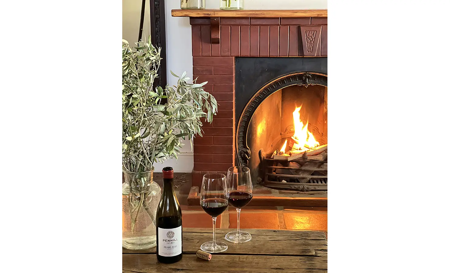 Perfect Hideaways: A wooden table set in front of a fireplace, adorned with a vase of olive branches, two glasses of red wine, and a bottle of Penhill wine. Penhill Manor House, Nuy Valley.