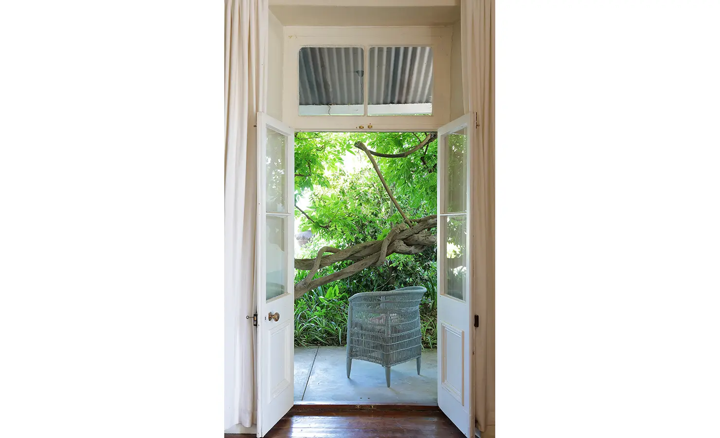 Perfect Hideaways: A serene garden view through open French doors, where a twisted tree frames a quiet corner with a woven chair, inviting peaceful contemplation. Staart Farm, Paarl.