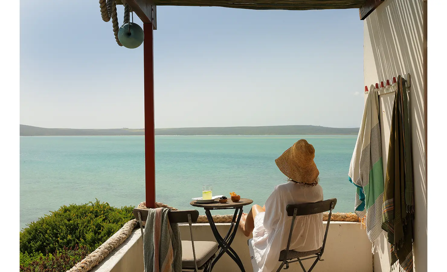 Perfect Hideaways: A woman in a straw hat enjoys a quiet moment at a bistro table with a drink and snacks, gazing out over tranquil turquoise waters from a shaded terrace. Churchhaven, Fynbos.