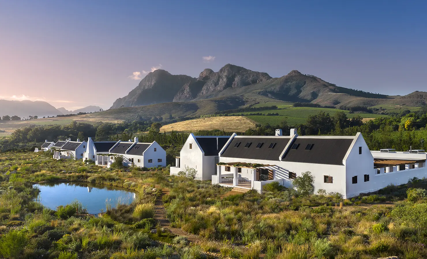 Perfect Hideaways: A serene landscape of Cape Dutch-style cottages surrounded by lush greenery, rolling vineyards, and towering mountain peaks at sunrise. Babylonstoren, Franschhoek.