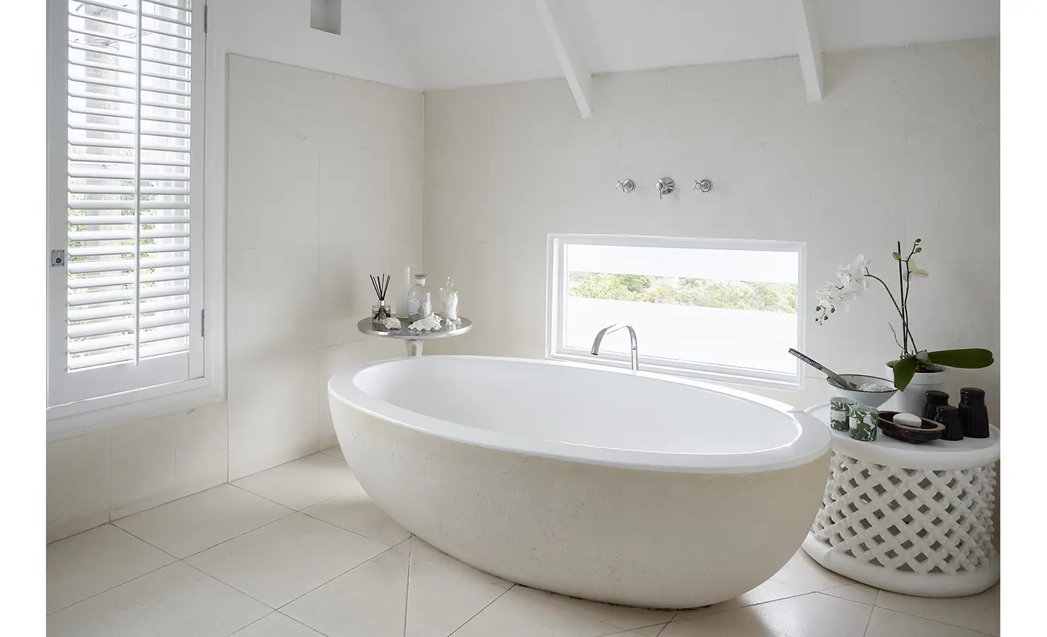 Perfect Hideaways: A sculptural freestanding bathtub takes centre stage in a light-filled bathroom with creamy tiles, shuttered windows, and delicate styling that includes orchids, candles, and bath essentials. Noordhoek Beach House, Noordhoek.
