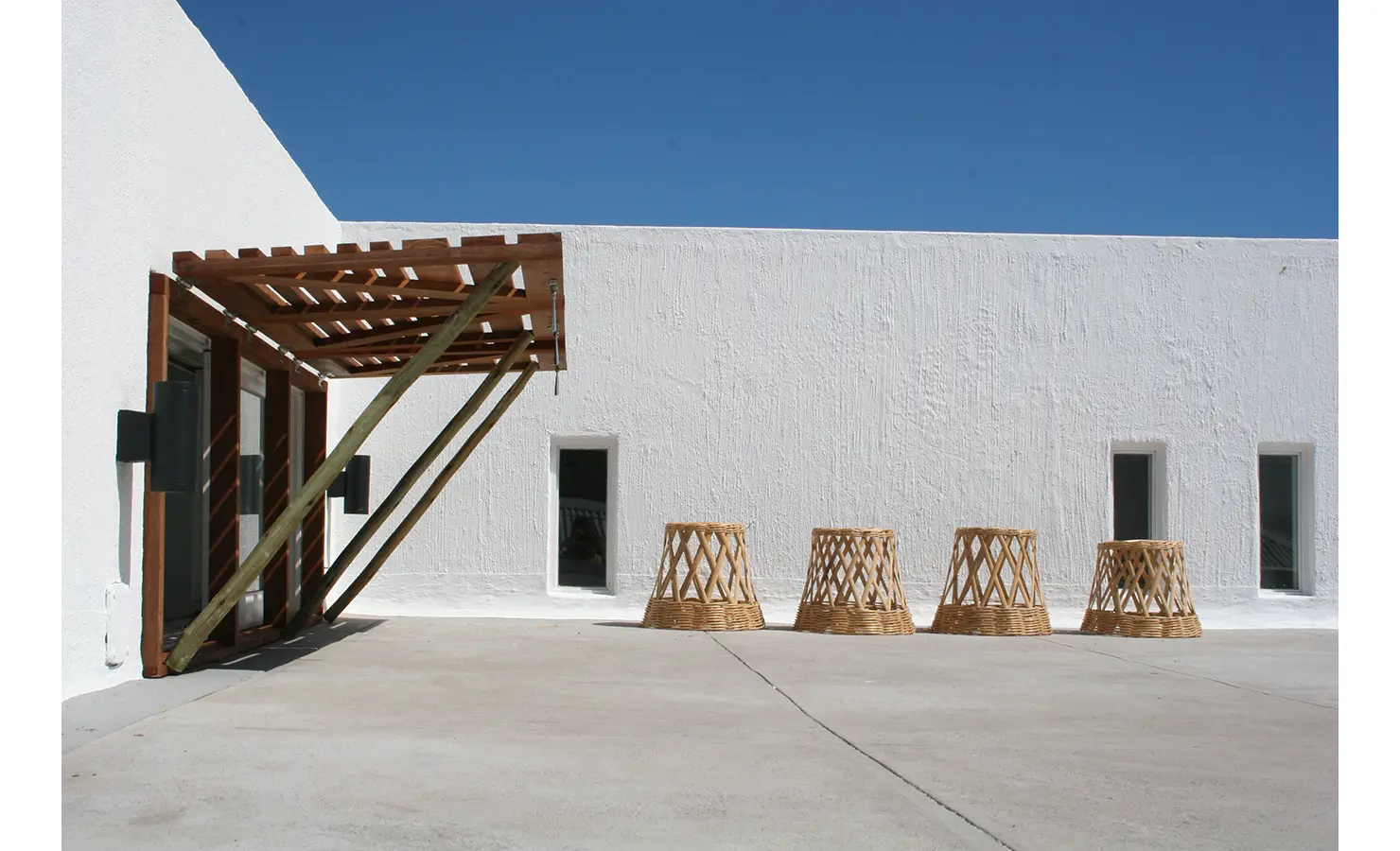 Perfect Hideaways: A striking white-walled courtyard with a simple wooden pergola and woven rattan stools set against a brilliant blue sky, creating a minimalist coastal aesthetic. Blue Lagoon, Langebaan.