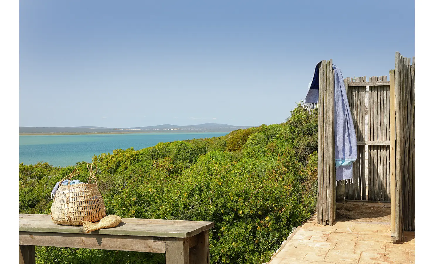 Perfect Hideaways: A rustic outdoor shower and weathered timber table overlook a stretch of lush green fynbos and the shimmering lagoon beyond under a clear blue sky. Churchhaven, Fynbos.