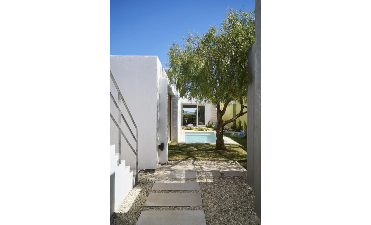 Perfect Hideaways: A tranquil garden pathway leading to a serene pool area, shaded by a lush tree, with modern white walls and stepping stones enhancing the natural setting. Blue Lagoon, Langebaan.