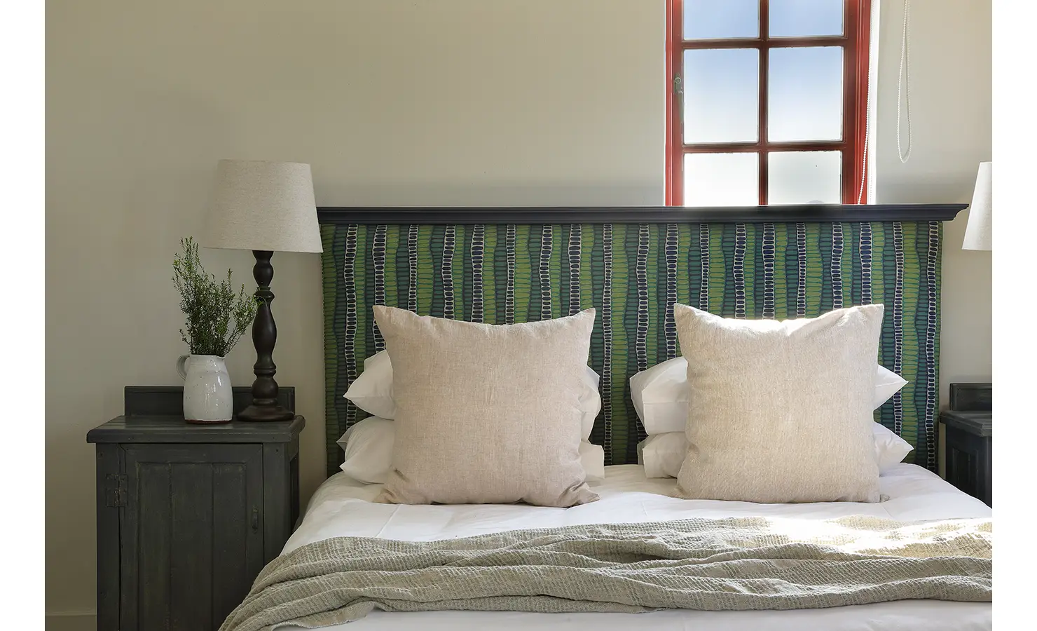 Perfect Hideaways: A cosy bedroom scene with a woven green and navy headboard, large linen cushions, and soft lighting streaming through a red-framed window. Churchhaven, Fynbos.
