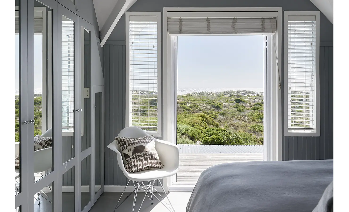 Perfect Hideaways: A serene grey-toned bedroom with mirrored wardrobes and a modern white armchair, offering uninterrupted views of indigenous fynbos and distant sand dunes. Noordhoek Beach House, Noordhoek.