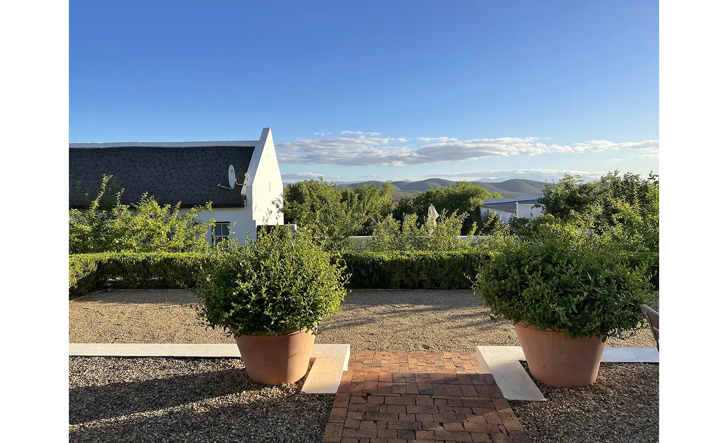 Perfect Hideaways: A picturesque courtyard framed by large terracotta pots with lush greenery, offering a breathtaking view of rolling hills and Cape Dutch architecture against a clear blue sky. Penhill Fruit Barn, Nuy Valley.