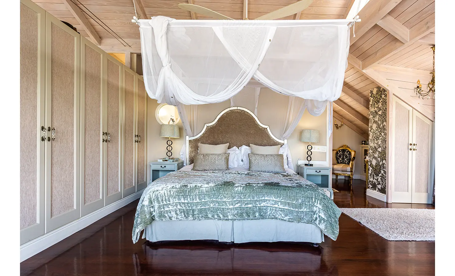 Perfect Hideaways: A luxurious bedroom featuring a grand upholstered bed, sheer canopy draping, and soft metallic accents, all beneath an exposed beam ceiling. Dolphin Cottage, Camps Bay.