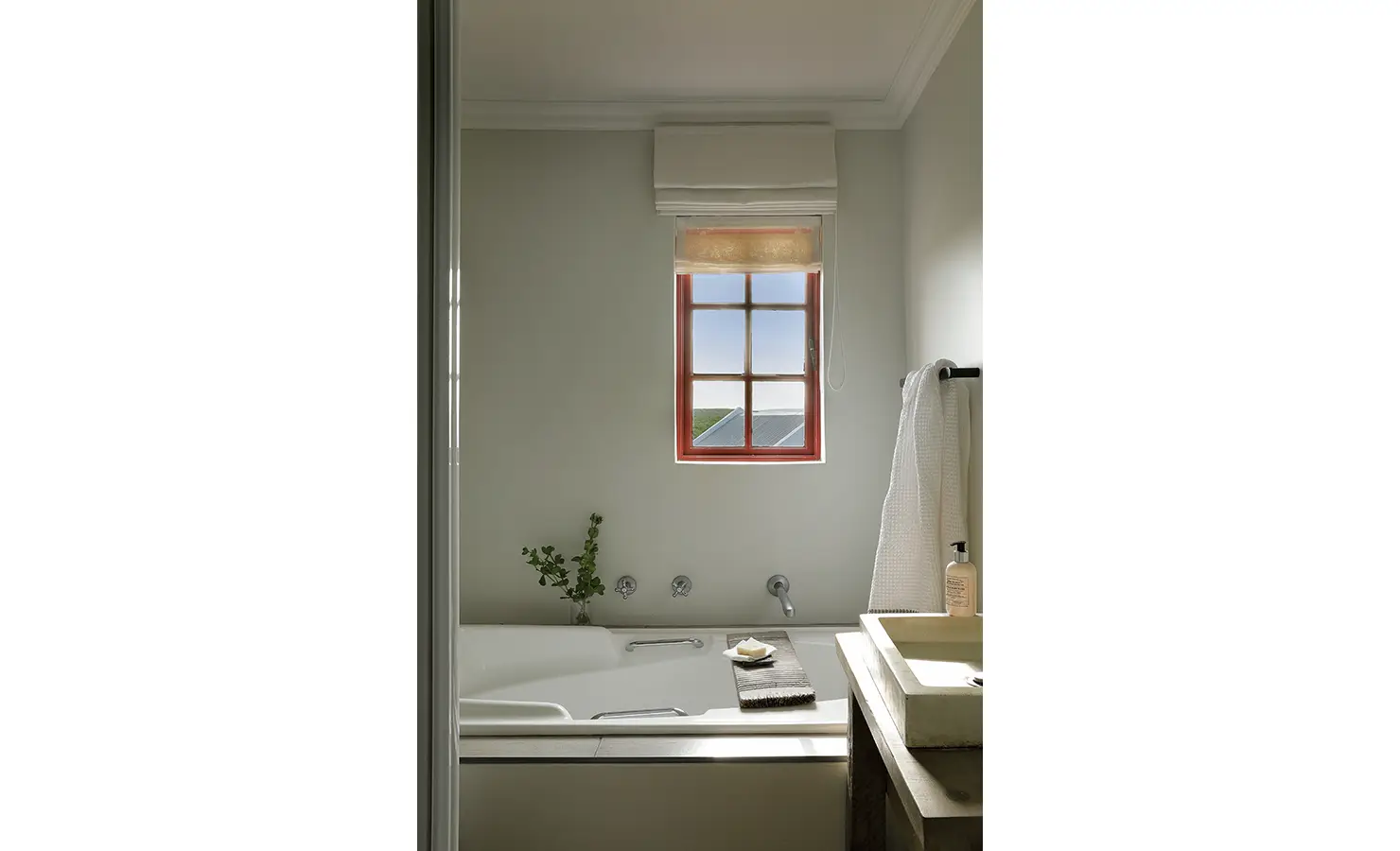 Perfect Hideaways: A serene bathroom with a deep tub and wooden vanity, where sunlight filters through a red-framed window above a zinc roofline. Churchhaven, Fynbos.
