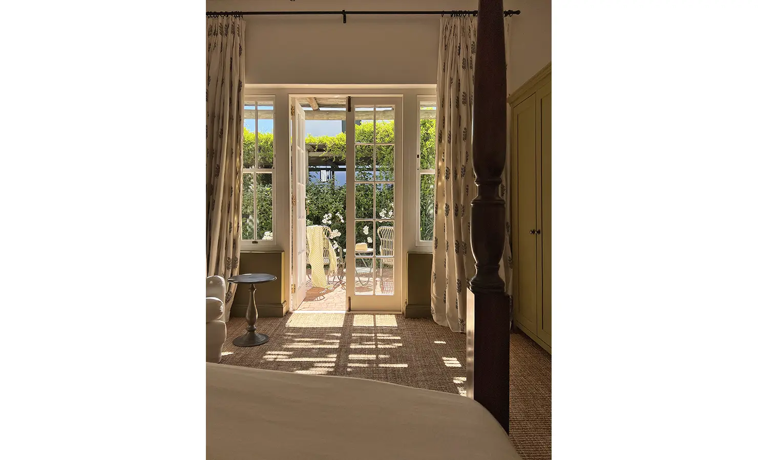 Perfect Hideaways: A tranquil bedroom view with French doors opening to a patio surrounded by vibrant greenery and dappled sunlight. Penhill Manor House, Nuy Valley.