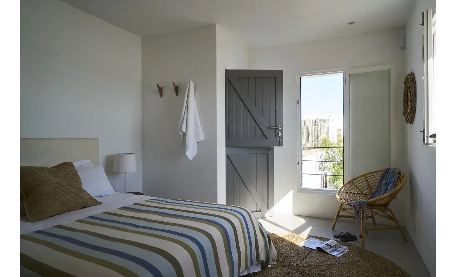 Perfect Hideaways: A cosy bedroom with a striped bedspread, a rustic wooden chair, and a Dutch-style stable door opening onto a bright outdoor space. Blue Lagoon, Langebaan.