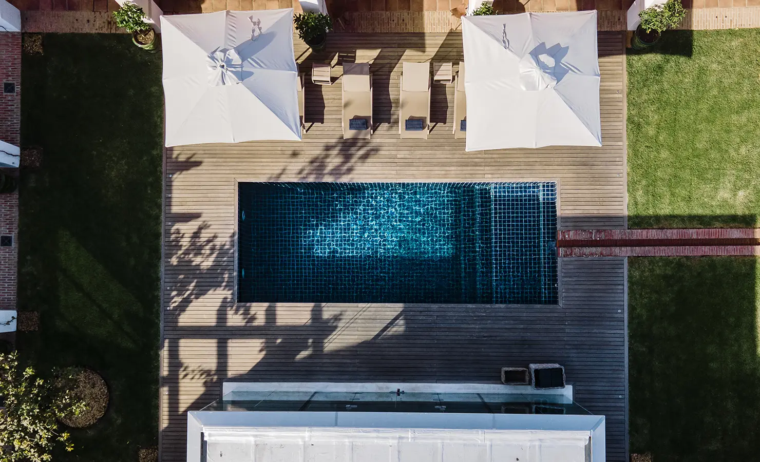 Perfect Hideaways: A striking aerial view of a modern swimming pool, wooden deck, and sun loungers, surrounded by lush green lawns and shaded seating areas. Babylonstoren, Franschhoek.
