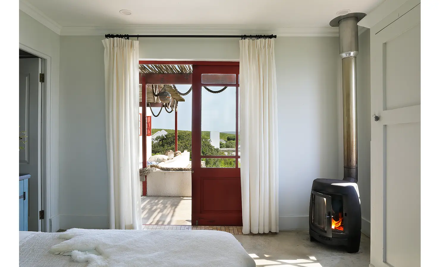 Perfect Hideaways: A snug bedroom features a fireplace, gauzy white curtains, and French doors that open onto a shaded veranda overlooking green coastal hills. Churchhaven, Fynbos.