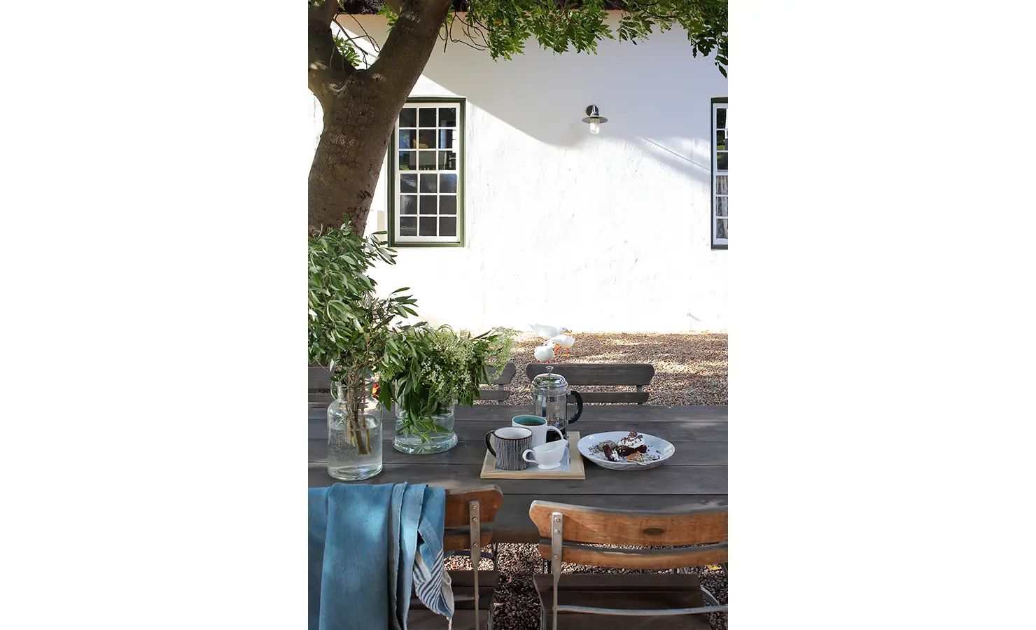 Perfect Hideaways: An outdoor dining table under the shade of a tree, set with cups, a French press, and a plate of treats, in front of a white-walled house. Penhill Manor House, Nuy Valley.