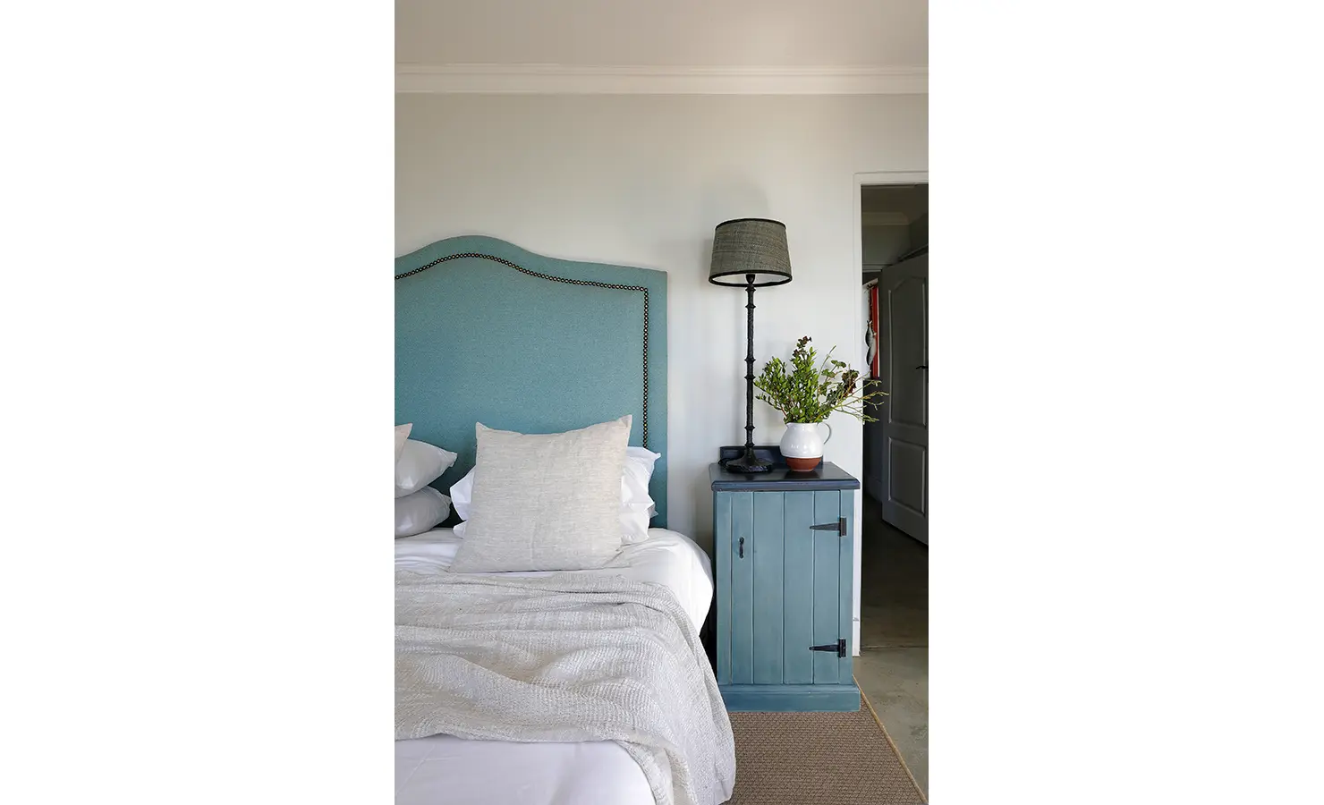 Perfect Hideaways: A refined bedroom corner with a curved teal headboard, textured bedding, and a vintage-style bedside table topped with a ceramic vase and botanical arrangement. Churchhaven, Fynbos.