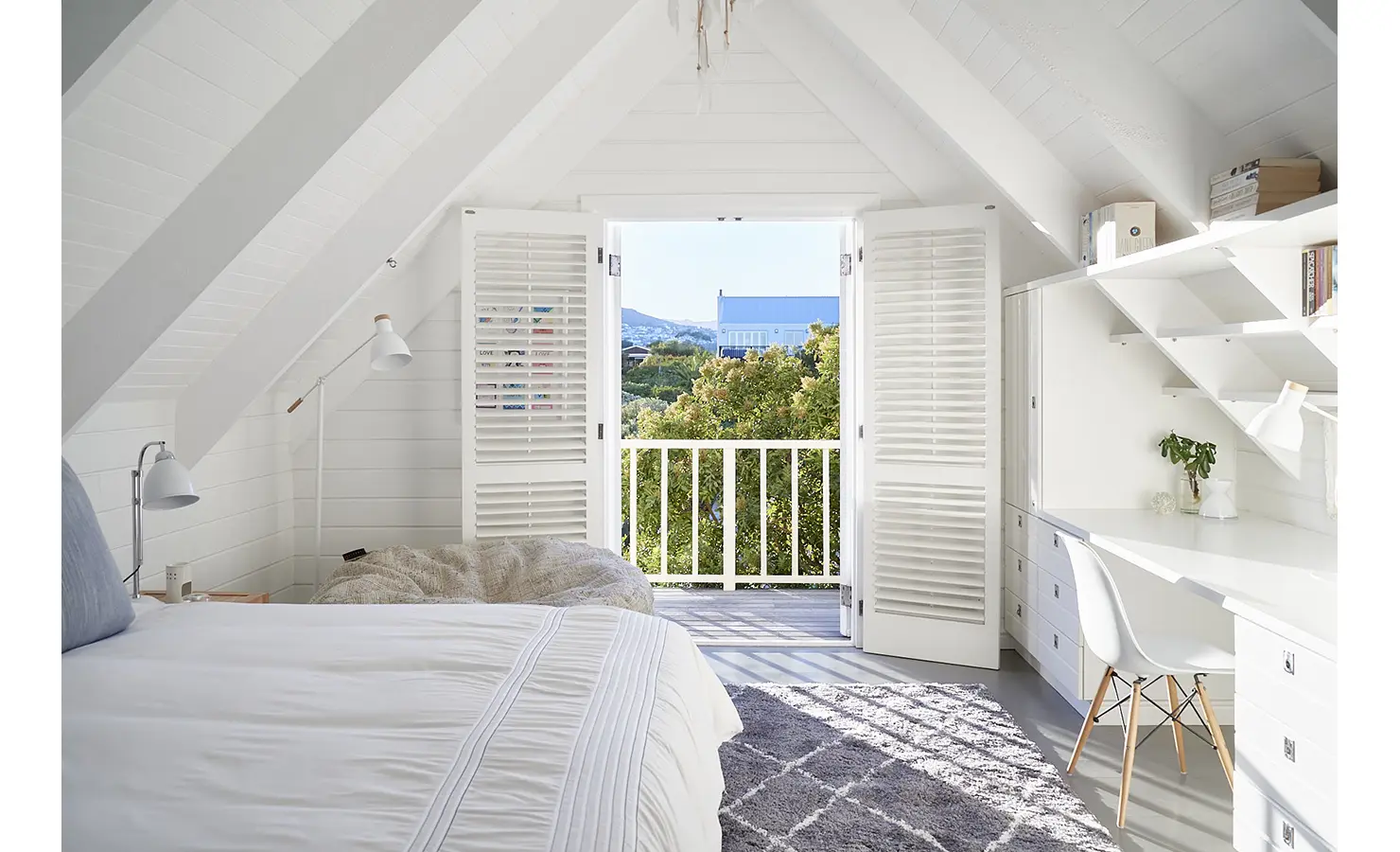 Perfect Hideaways: A light and airy loft-style bedroom with a sloped ceiling, white beams, built-in workspace, and French doors leading to a treetop balcony with leafy views. Noordhoek Beach House, Noordhoek.