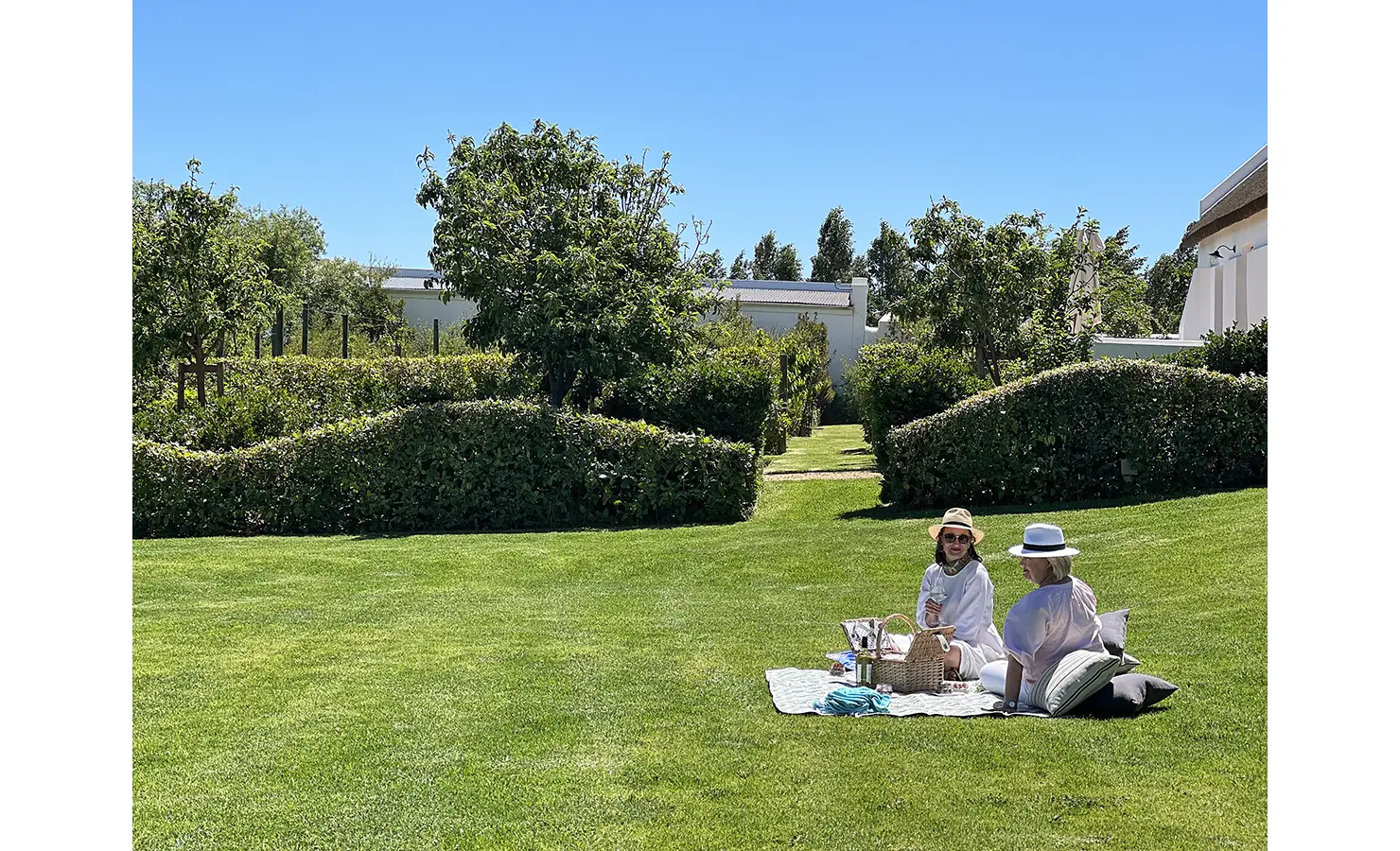 Perfect Hideaways: Two people enjoying a picnic on a lush green lawn, surrounded by manicured hedges and trees under a bright blue sky. Penhill Manor House, Nuy Valley.