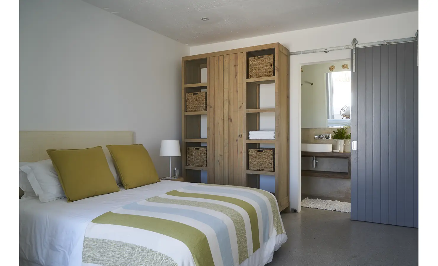 Perfect Hideaways: A beautifully designed bedroom with a soft colour palette, a rustic wooden wardrobe, and an en-suite bathroom accessed via a sliding barn-style door. Blue Lagoon, Langebaan.