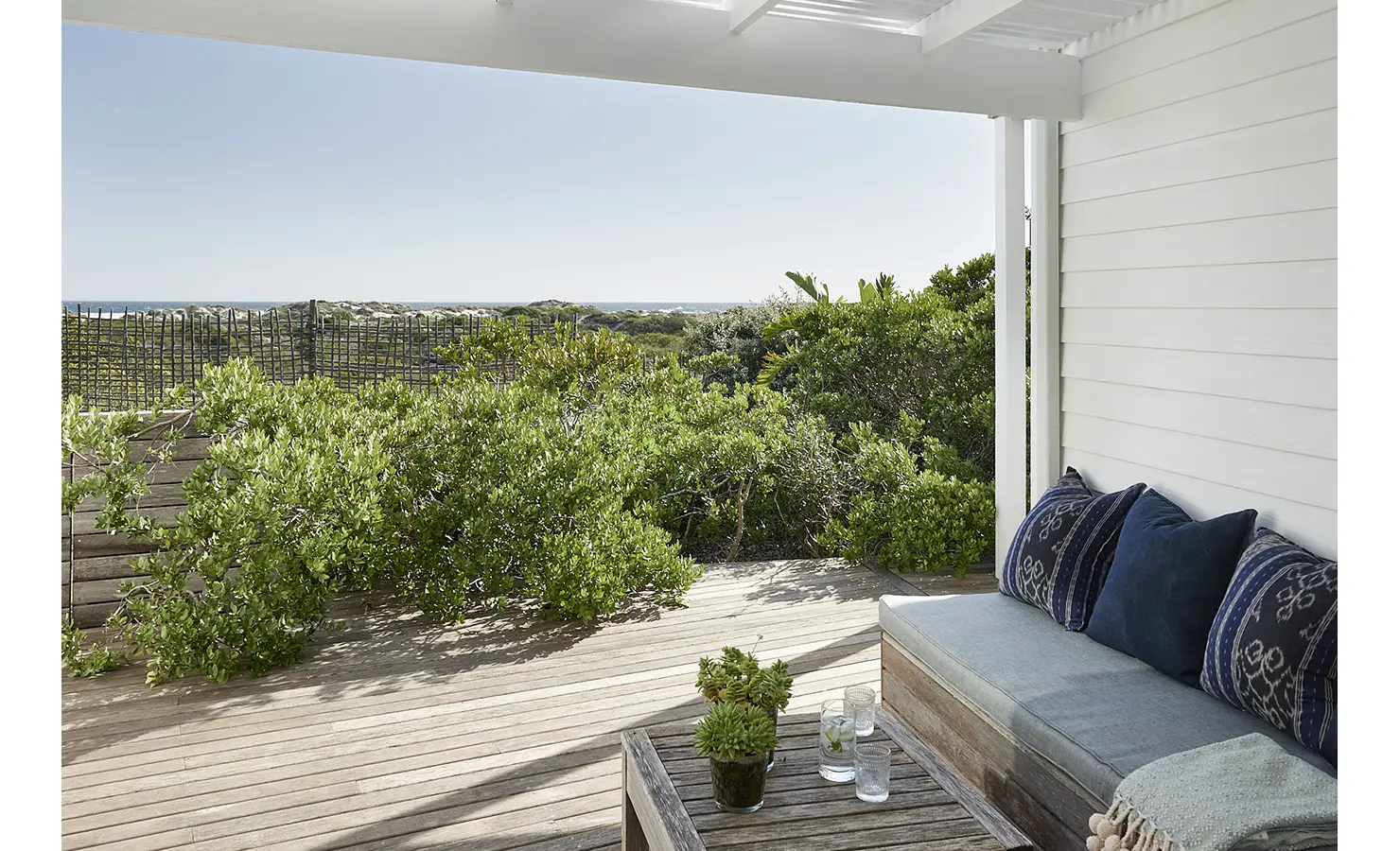 Perfect Hideaways: A peaceful covered veranda with a built-in daybed, deep indigo cushions, and a rustic wooden table, overlooking a deck framed by indigenous shrubs and views of the ocean beyond. Noordhoek Beach House, Noordhoek.