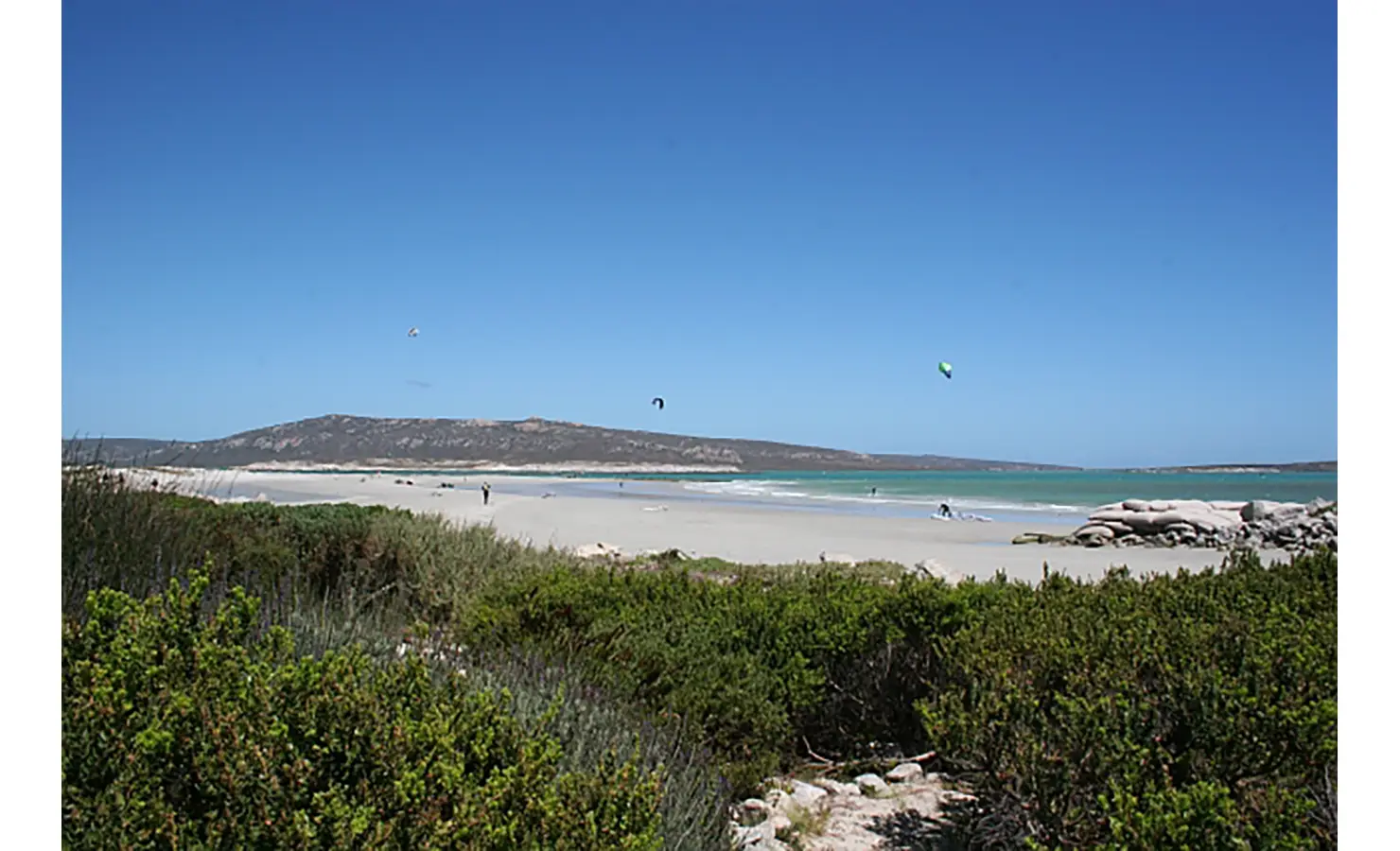 Perfect Hideaways: A breathtaking view of the pristine beach with soft white sand, turquoise waters, and kite surfers gliding in the distance under a clear blue sky. Blue Lagoon, Langebaan.
