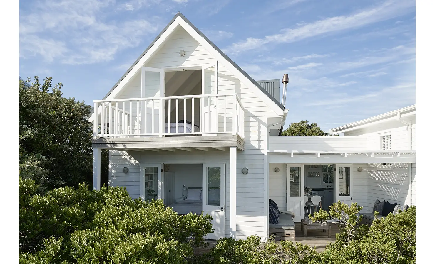Perfect Hideaways: A charming white timber guest cottage with a gabled roof and balcony, surrounded by lush greenery, featuring multiple French doors opening onto relaxed outdoor living areas. Noordhoek Beach House, Noordhoek.