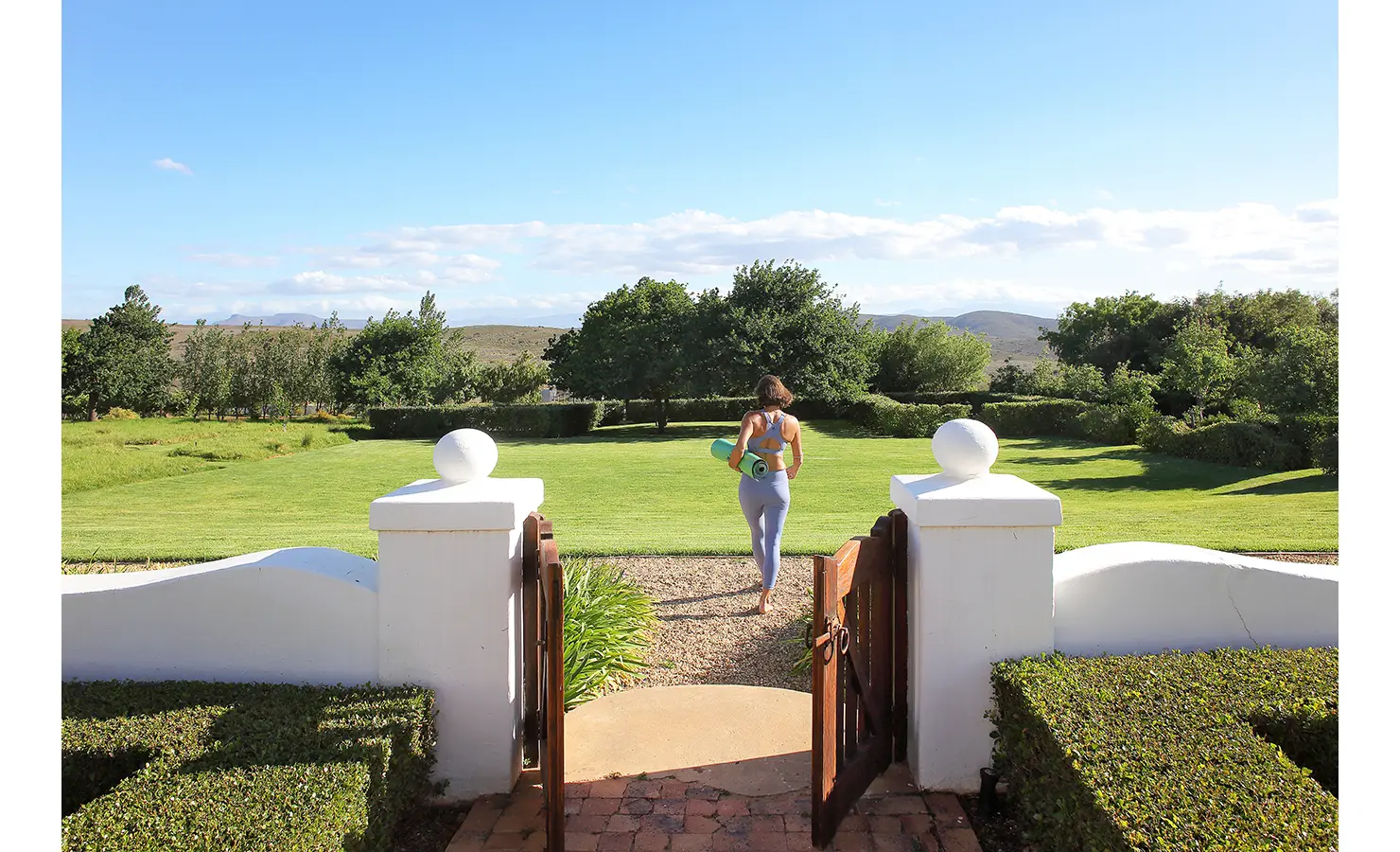 Perfect Hideaways: A woman in activewear walking through a gate into a lush green garden, carrying a yoga mat, with rolling hills in the distance. Penhill Manor House, Nuy Valley.