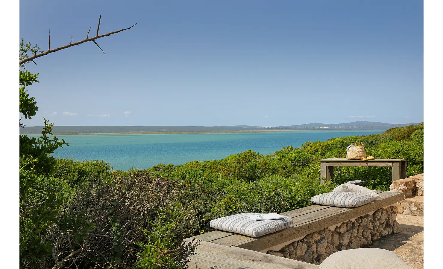 Perfect Hideaways: A serene outdoor bench with striped cushions overlooking the shimmering turquoise lagoon and lush greenery under a clear blue sky, Churchhaven, Fynbos.