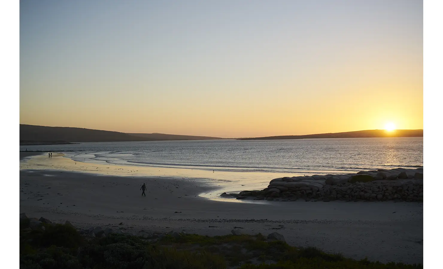 Perfect Hideaways: A peaceful shoreline at dusk, where the golden sun sets over calm waters, casting a soft reflection on the wet sand and rocky outcrops. Blue Lagoon, Langebaan.