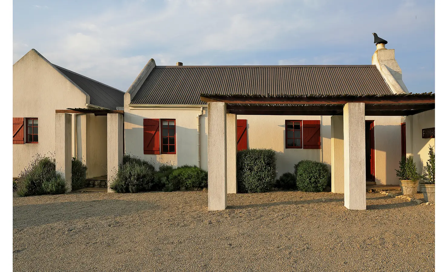 Perfect Hideaways: A charming whitewashed coastal cottage with red window shutters, pitched corrugated iron roof and a shaded veranda set against a warm golden light, Churchhaven, Fynbos.