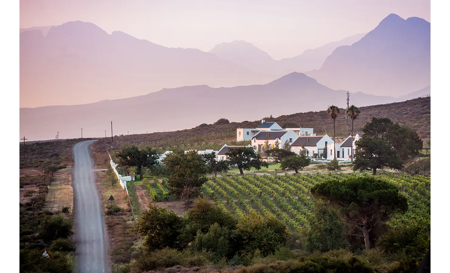 Perfect Hideaways: An aerial view of the estate surrounded by vineyards and open landscape, with majestic mountains in the distance at dusk. Penhill Manor House, Nuy Valley.