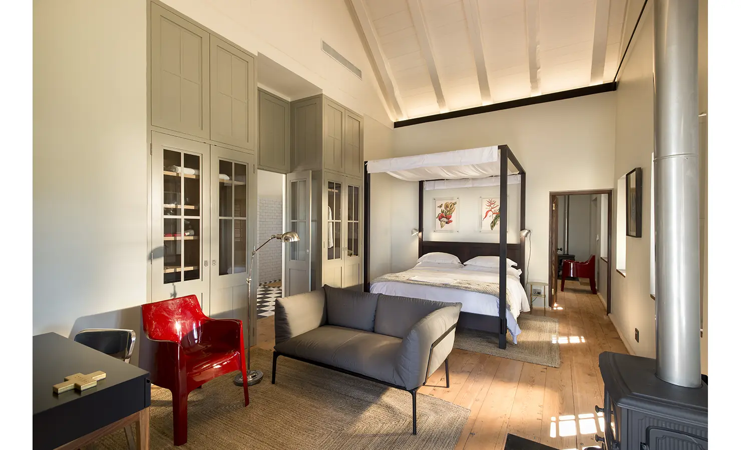 Perfect Hideaways: A spacious bedroom featuring a dark four-poster bed, muted sage cabinetry, and botanical prints, with an elegant en-suite bathroom visible in the background. Babylonstoren, Franschhoek.
