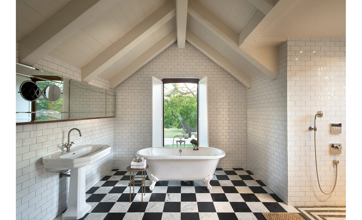 Perfect Hideaways: A light-filled bathroom with a freestanding clawfoot bathtub, checkerboard flooring, and large double doors opening onto a lush green courtyard. Babylonstoren, Franschhoek.