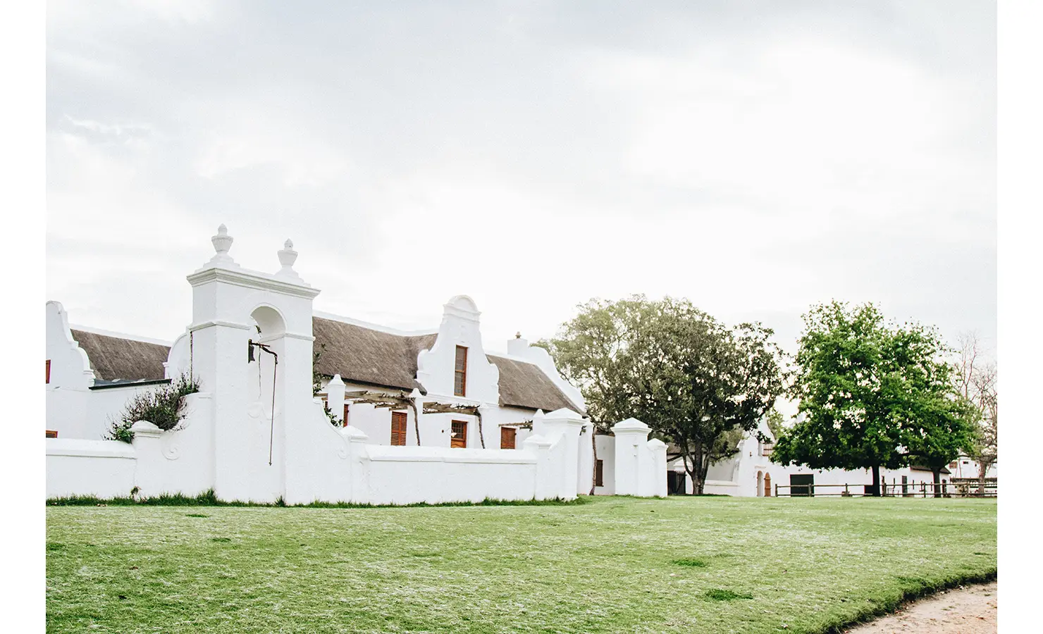 Perfect Hideaways: A grand Cape Dutch farmhouse with crisp white walls, thatched roofing, and wooden shutters, set against a soft, overcast sky. Babylonstoren, Franschhoek.