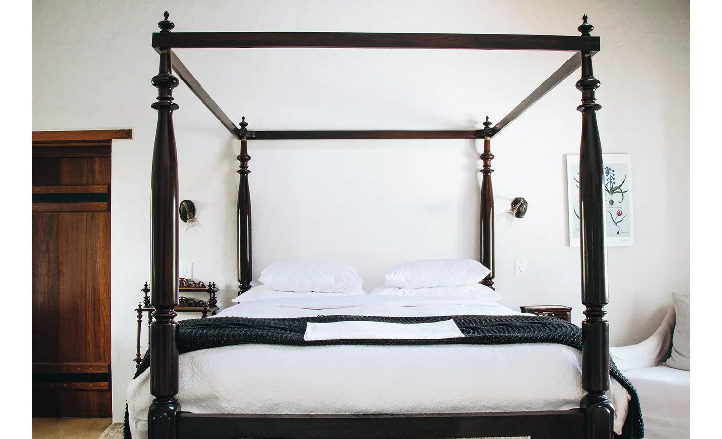 Perfect Hideaways: A striking dark wood four-poster bed with intricately carved posts, crisp white bedding, and botanical artwork adorning the walls in a serene bedroom. Babylonstoren, Franschhoek.