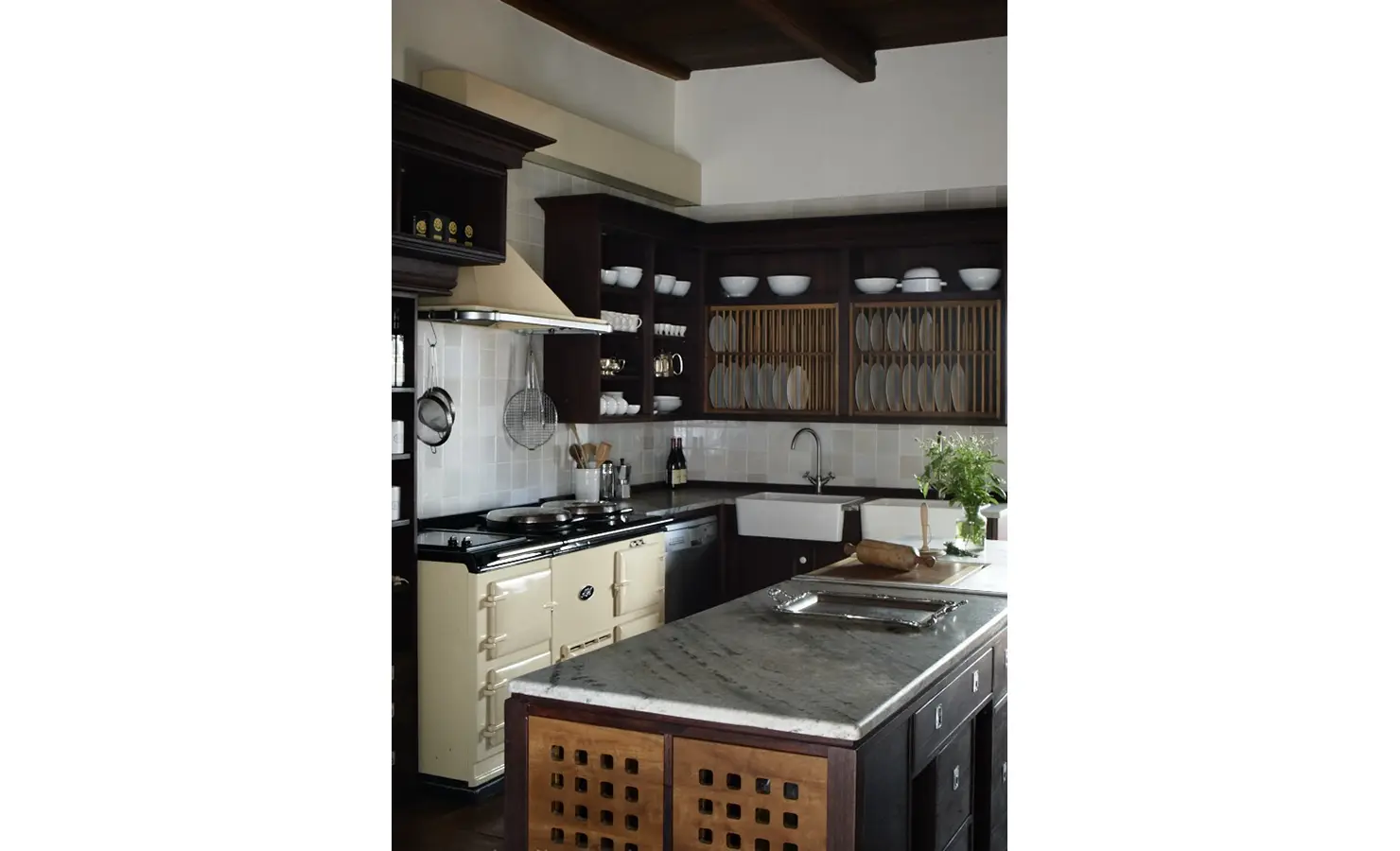 Perfect Hideaways: A traditional farmhouse kitchen with dark wooden cabinetry, a vintage cream AGA stove, and open shelving displaying rustic dishware and cookware. Babylonstoren, Franschhoek.