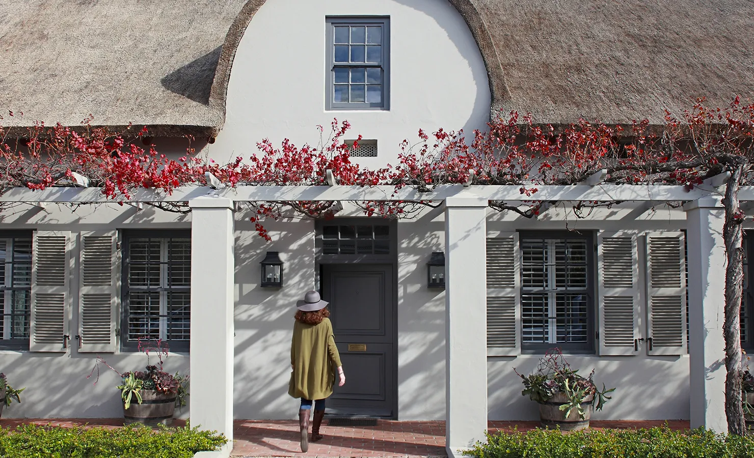 Lady walking towards thatch manor farm house with shutters and vine pergola. Koko House.