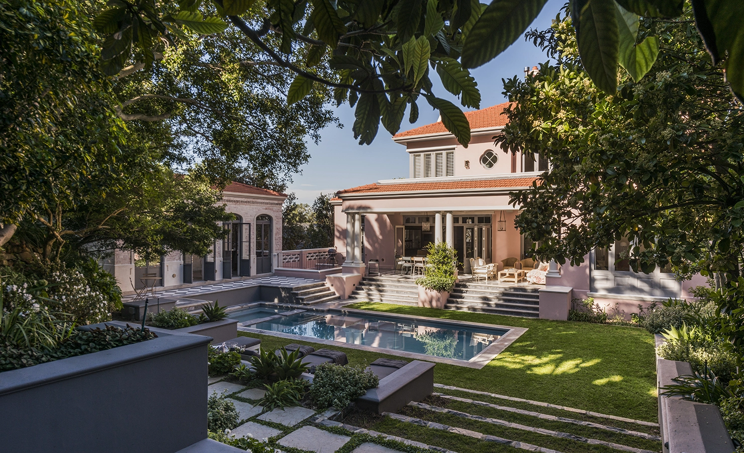 Perfect Hideaways: A serene garden sanctuary surrounds a shimmering rectangular pool, framed by manicured greenery and mature trees, leading up to a beautifully restored villa with blush pink walls, shuttered windows, and a classic red-tiled roof — evoking timeless elegance in a private city retreat. Hope House, Cape Town City, Oranjezicht.