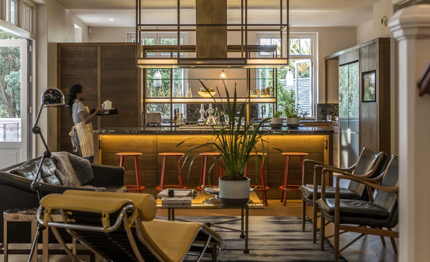 Perfect Hideaways: A warm, open-plan kitchen features wooden cabinetry, a glowing central island with bright red stools, and a glimpse of cosy, art-filled living space. Hope House, Cape Town City, Oranjezicht.