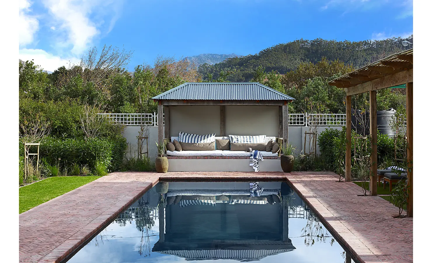 Outdoor swimming pool next to seated pergola surrounded by green trees. Koko House.