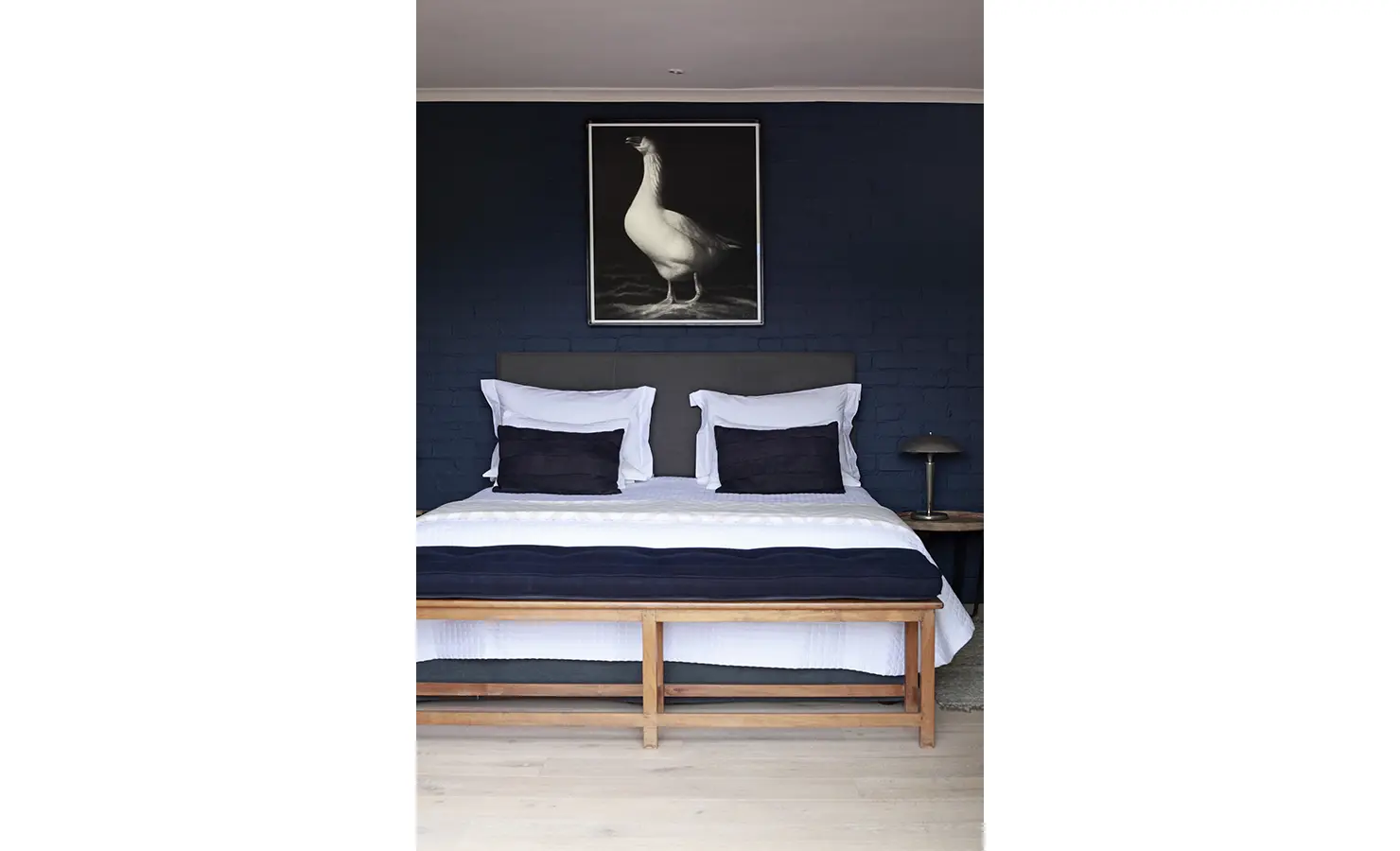 Dark blue painted bedroom wall with bed, wooden bedend and large black and white goose image. Koko House.