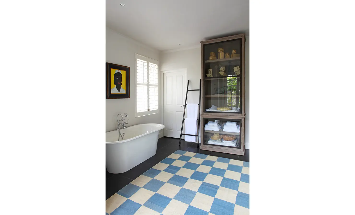 Free standing bath with blue and white checkered rug, bright yellow artwork and wooden display cabinet. Koko House.