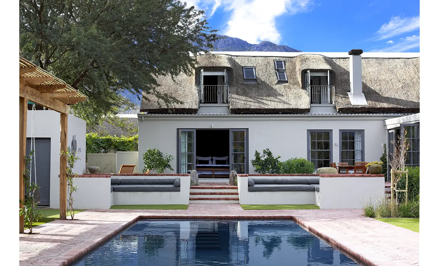 Outdoor swimming pool with backdrop of thatch manor farmhouse and patio area. Koko House.