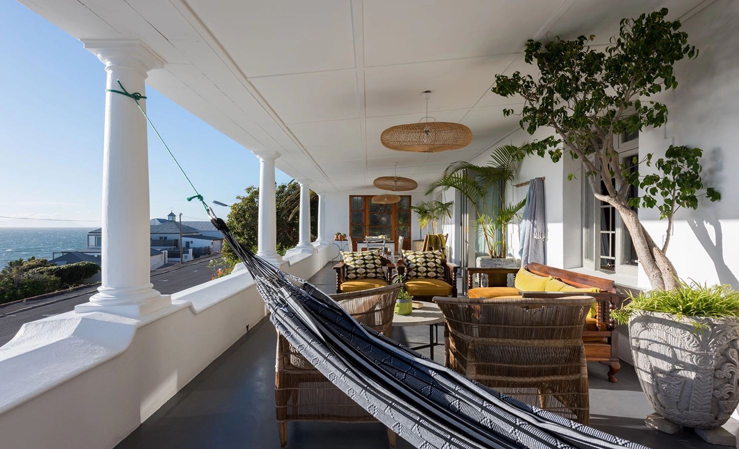 Perfect Hideaways: A breezy, colonial-style veranda overlooking the ocean, featuring white pillars, a woven hammock, and an inviting seating area with wicker chairs, patterned cushions, and tropical potted plants — ideal for laid-back coastal living. Blue Waters, Simons Town.