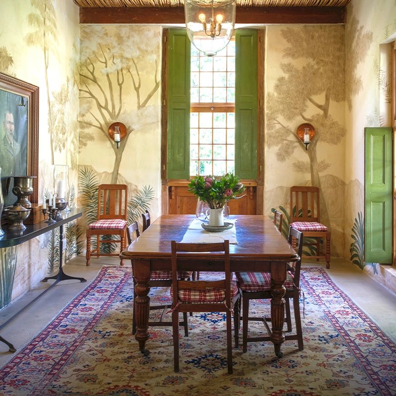 Perfect Hideaways: An elegant dining room with hand-painted botanical murals, antique wooden furniture, a Persian-style rug, and green shuttered windows that let in dappled natural light, creating a warm and nostalgic atmosphere. De Hoek, Barrydale.