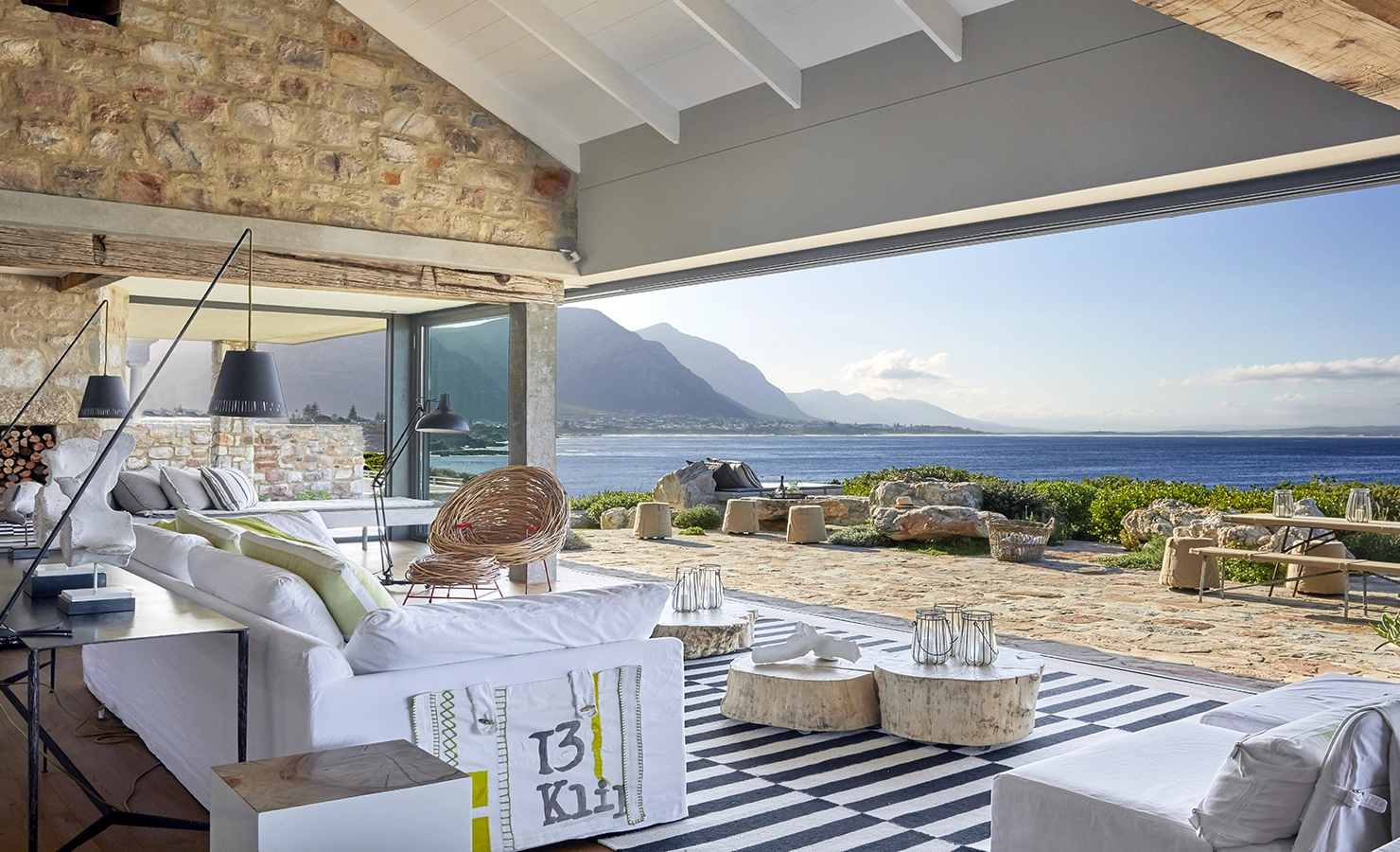 Perfect Hideaways: A light-filled open-plan living area with stone walls and exposed beams flows seamlessly onto a spacious terrace, offering uninterrupted views of Walker Bay and the dramatic mountains beyond. Stone House, Hermanus.