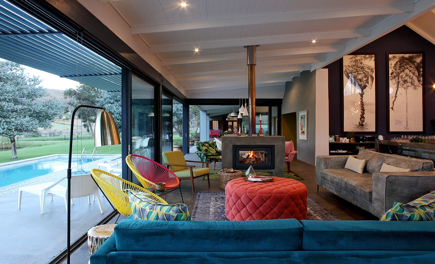 Perfect Hideaways: A vibrant and eclectic living space features bold furniture in rich textures and jewel tones, centred around a contemporary fireplace, with floor-to-ceiling glass doors opening to a poolside terrace and views of the Karoo landscape beyond. 22 Degrees East, Prince Albert.