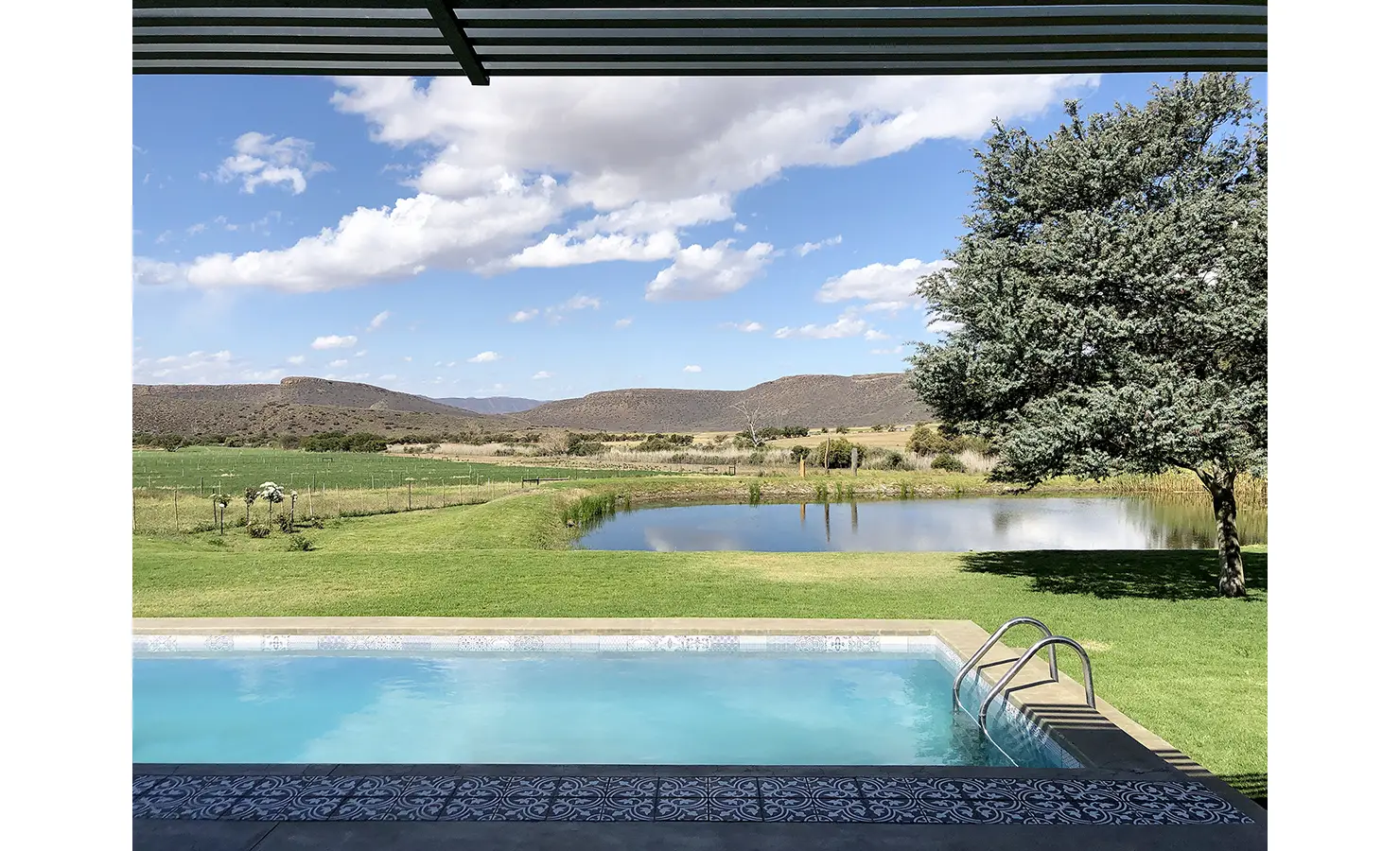 Perfect Hideaways: A serene outdoor setting with a turquoise swimming pool and manicured lawn, overlooking a peaceful dam and the rugged Karoo mountains under a wide blue sky. 22 Degrees East, Prince Albert.
