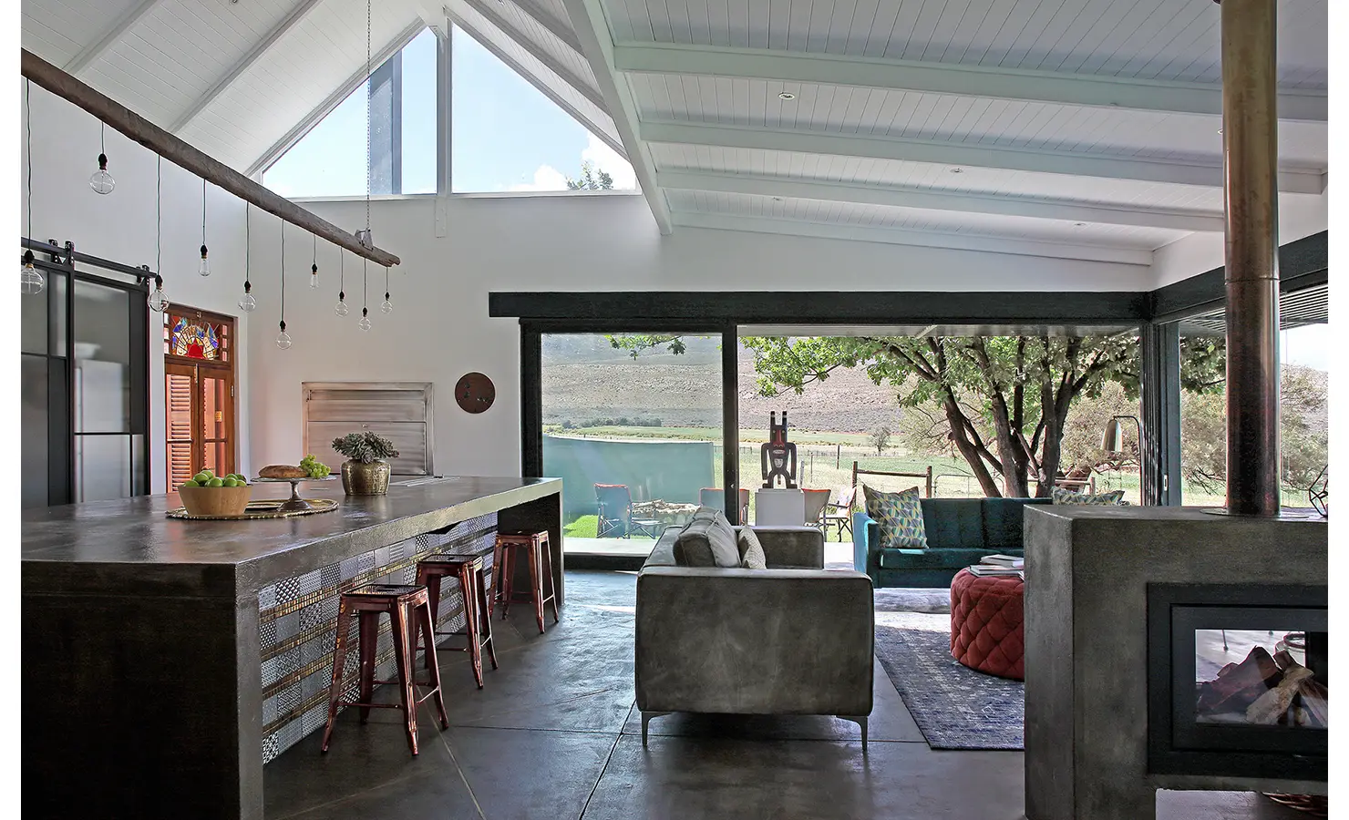 Perfect Hideaways: A spacious open-plan living area blends industrial chic with rustic charm, featuring polished concrete finishes, large windows framing mountain views, and a striking fireplace. 22 Degrees East, Prince Albert.