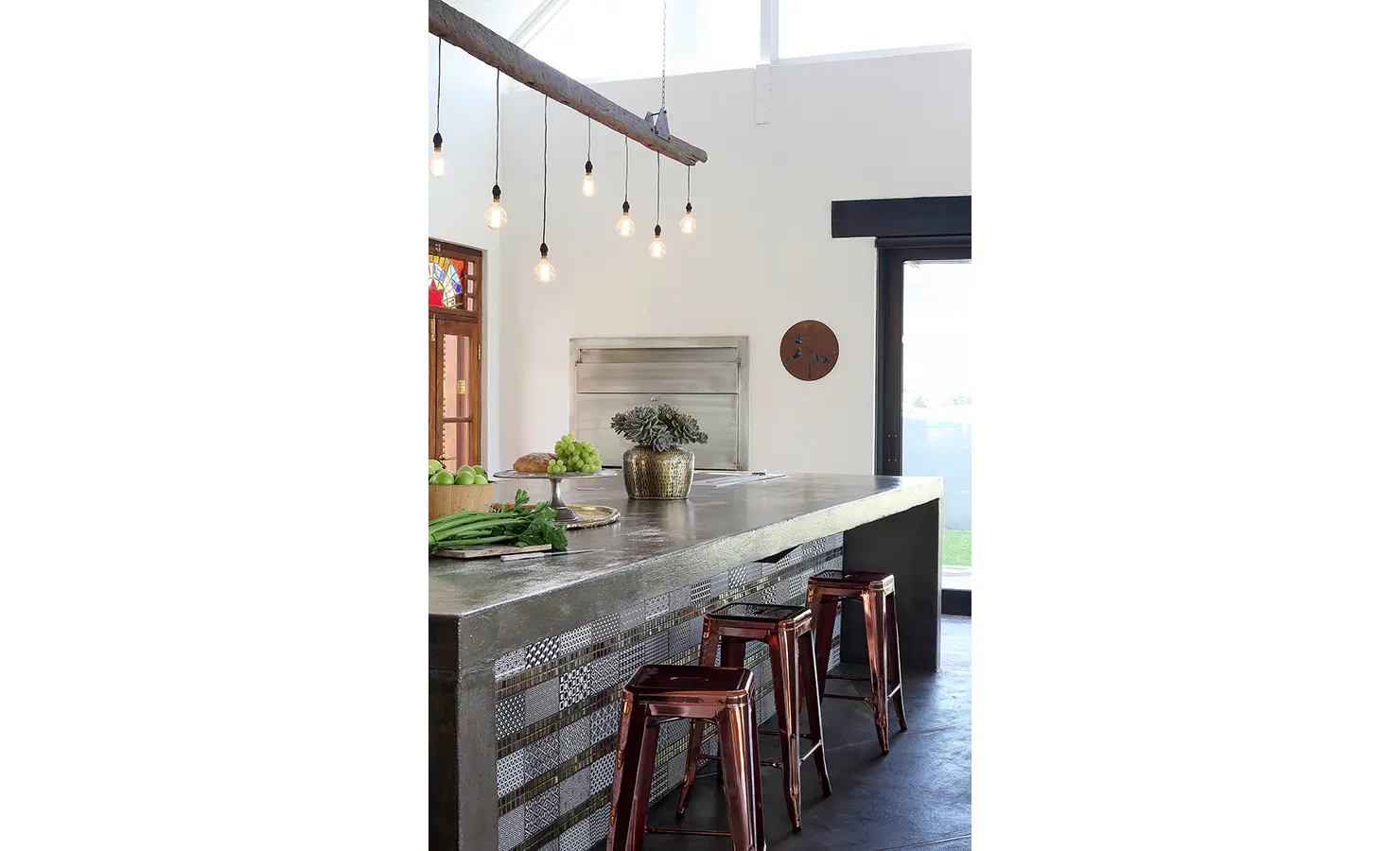 Perfect Hideaways: A raw concrete kitchen island with copper-toned bar stools is illuminated by exposed bulbs hanging from a rustic wooden beam, combining industrial elegance with warmth. 22 Degrees East, Prince Albert.
