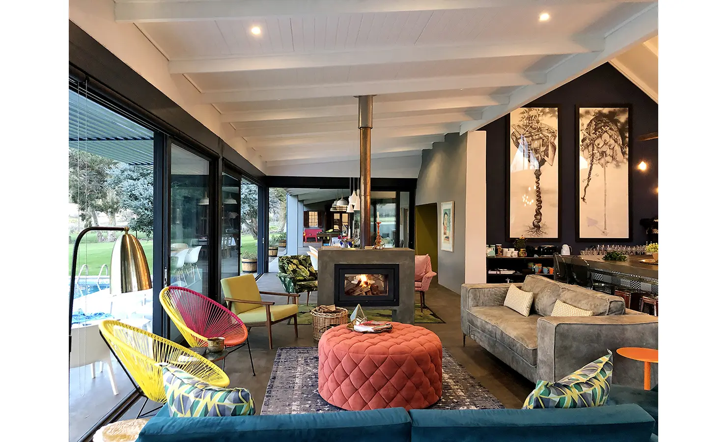Perfect Hideaways: A vibrant and eclectic living space features bold furniture in rich textures and jewel tones, centred around a contemporary fireplace, with floor-to-ceiling glass doors opening to a poolside terrace and views of the Karoo landscape beyond. 22 Degrees East, Prince Albert.